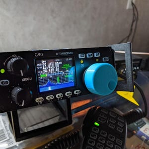 May include: A black Xiegu G90 HF transceiver featuring a digital display showing 28.410.00 MHz, two black control knobs, a bright blue tuning dial, and several labeled buttons. The device is mounted in a black metal frame with a handheld microphone.