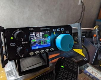 Może przedstawiać: Czarny radiotelefon Xiegu G90 HF z cyfrowym wyświetlaczem pokazującym 28.410.00 MHz, dwoma czarnymi pokrętłami, jaskrawoniebieskim pokrętłem strojenia i kilkoma opisanymi przyciskami. Urządzenie jest zamontowane w czarnej metalowej ramie.
