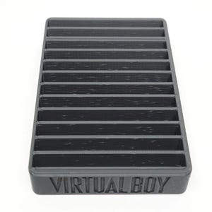 May include: A rectangular black plastic storage rack featuring twelve horizontal slots for game cartridges. The front edge of the base is embossed with the text VIRTUAL BOY in capital letters. The object has a textured finish and a grid-like design.