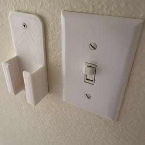 May include: A white plastic wall-mounted holder with a rectangular backplate and a U-shaped cradle, secured by a single screw. Positioned next to it is a standard white rectangular light switch plate featuring a single toggle switch and two screws.
