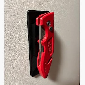 May include: A red Milwaukee Fastback utility knife mounted on a black rectangular magnetic holder. The utility knife features a metallic blade section and the brand name Milwaukee embossed on the side, secured to the black mount.