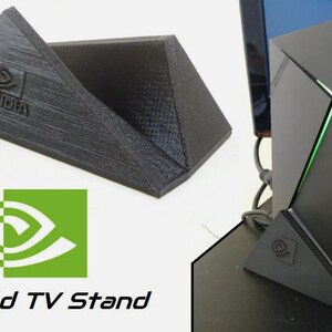 May include: A black 3D-printed triangular stand for an NVIDIA Shield TV device. The stand features a textured surface and an embossed NVIDIA logo on the front. It is designed to hold the console in an upright vertical position for display.