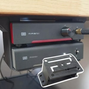 May include: A black under-desk mounting bracket for audio equipment, shown alongside a Schiit Magni headphone amplifier and a Schiit Modi digital-to-analog converter. The bracket features a rectangular frame with screw holes for installation.