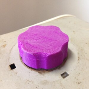 May include: A bright purple, six-lobed knob with a textured, layered surface finish. The knob is circular with a scalloped edge and is mounted onto a light gray, weathered base that features two small, square-shaped cutouts on opposite sides.