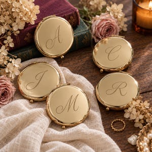 May include: Four round gold-toned metal compact mirrors, each engraved with a single cursive letter: A, C, J, M, or R. The mirrors are arranged on a textured cream-coloured fabric, surrounded by dried flowers, vintage books, and a small gold ring.