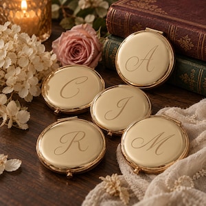 May include: Five round, gold-toned metal compact mirrors, each featuring a single engraved script letter on the lid. The letters A, C, J, M, and R are clearly visible on the polished surfaces of these hinged, circular vanity accessories.