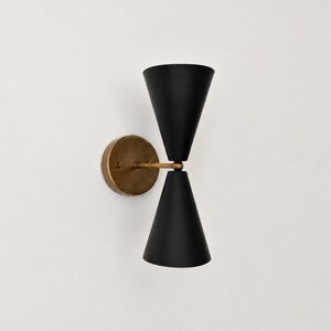 May include: A wall-mounted light fixture featuring two matte black conical shades arranged in a vertical hourglass configuration, connected by a central brass-toned arm to a circular brass-toned mounting plate that attaches to the wall.