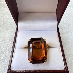 May include: A gold ring featuring a large, rectangular, emerald-cut amber-colored gemstone held by four prongs. The ring is displayed inside a dark burgundy jewelry box with a white interior lining, showcasing the stone's deep orange hue.