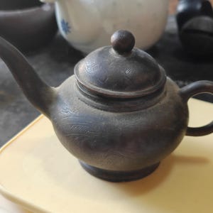 May include: A dark brown, metallic teapot with a rounded body, a curved spout, and a matching lid featuring a small knob. The surface of the teapot is decorated with fine, etched line patterns that wrap around the exterior of the vessel.