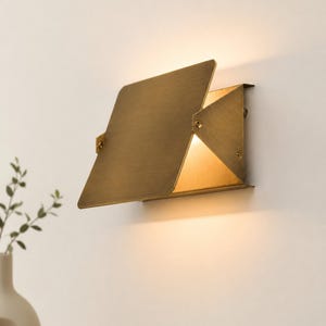 May include: A wall-mounted light fixture with a brushed brass finish, featuring a rectangular metal plate angled outward. The design includes triangular side panels and exposed screws, emitting a warm, soft glow from behind the plate.