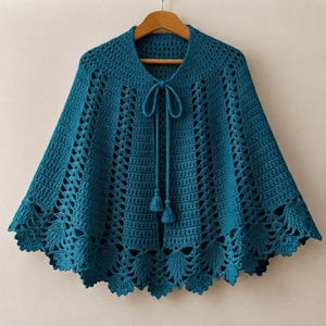 Op de afbeelding: Een blauwgroene gehaakte capelet met een verticaal steekpatroon met opengewerkte details, een ronde halslijn en een sluiting aan de voorzijde met twee bijpassende kwastjes. De zoom is afgewerkt met een geschulpte rand en hangt aan een kledinghanger.