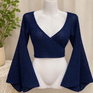 May include: A dark navy blue long-sleeve crop top featuring a wrap-style front with a deep V-neckline. The garment is constructed from a crocheted fabric with an open-knit texture and includes wide, flared bell sleeves that extend from the shoulders.