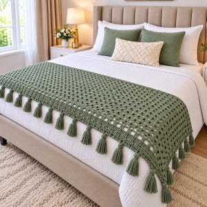 May include: A sage green crocheted bed runner featuring a repeating open-work square pattern and decorative tassels along the edges. The runner is draped over a white bedspread, complemented by two sage green pillows and a cream-colored lumbar pillow.