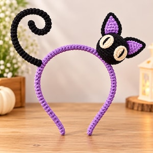 May include: A purple crochet headband featuring a black cat head with large white eyes and purple inner ears on the right side, and a black coiled cat tail extending from the left side. The entire structure is covered in textured yarn.