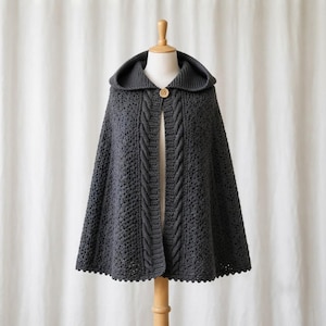 Op de afbeelding: Een donkergrijze cape met capuchon en een ingewikkeld opengewerkt haakpatroon over het hele lichaam. Het kledingstuk heeft een dikke, verticale kabelgebreide rand langs de opening aan de voorzijde en wordt bij de hals gesloten met een houten knoop.