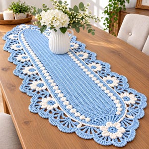 Può includere: Un runner da tavola ovale all'uncinetto con un centro azzurro a punto lavorato, bordato da un bordo decorativo smerlato composto da motivi floreali bianchi e dettagli a ventaglio azzurri, creando un design dettagliato simile al pizzo.