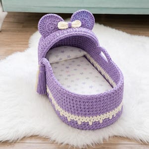 Puede incluir: Un moisés para muñecas de ganchillo color lavanda con una capota que tiene dos orejas redondeadas y un pequeño lazo crema. El moisés incluye una borla crema a juego en el lateral, una almohada blanca y un colchón blanco con estrellas grises.