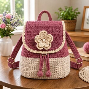 May include: A two-toned crochet backpack featuring a deep magenta upper section and a cream-colored lower section. The flap is adorned with a large, layered cream crochet flower with a central pearl bead and a cream border trim.