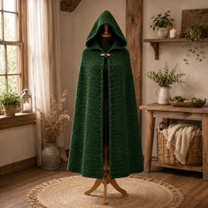 Op de afbeelding: Een donker bosgroene gehaakte cape met capuchon, voorzien van een gestructureerd steekpatroon en een enkele houten knevelsluiting bij de hals. Het kledingstuk wordt getoond op een paspop en heeft een lang ontwerp met een puntige capuchon.