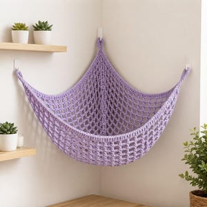 May include: A light purple crochet corner hammock featuring an open diamond-shaped mesh pattern. The triangular storage net is suspended by three loops attached to white wall hooks, creating a hanging storage space for various small items.