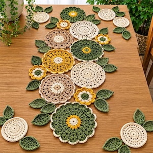 May include: A crochet table runner composed of various circular motifs in cream, beige, mustard yellow, and dark green. The design features intricate floral-inspired patterns and individual crocheted leaves arranged in a decorative cluster.