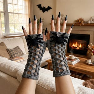 May include: A pair of gray fingerless gloves featuring a crocheted web pattern and a black spider appliqué on the back of each hand. The gloves extend from the knuckles to the mid-forearm and are crafted with a textured, open-work yarn design.