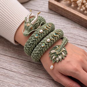May include: A sage green crocheted wrap bracelet featuring a dragon head with white horns, embedded moon phase charms, and a leaf-shaped charm at the end. The bracelet is accented with small white pearls and gold-toned metal details throughout.