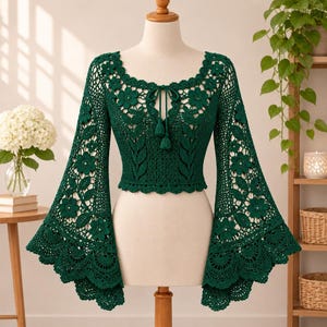 Puede incluir: Un top de crochet de manga larga en color verde oscuro con un patrón de flores y hojas. La prenda presenta un escote redondo con un cierre de lazo central que termina en dos borlas y mangas anchas acampanadas con borde festoneado.