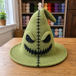 May include: A light green crocheted hat featuring a pointed top and a wide brim. The hat displays a dark black stitched pattern down the center, two dark black triangular eye shapes, and a wide, jagged black stitched mouth across the front.