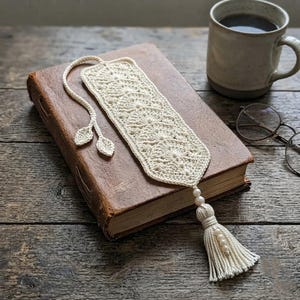 May include: An off-white crochet bookmark featuring a repeating fan-like pattern, a long cord ending in two small leaf-shaped accents, and a bottom tassel adorned with four small white beads, resting on a thick, weathered brown hardcover book.