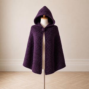 May include: A deep purple hooded cape crafted from thick, chunky yarn with a prominent crochet texture. The garment features a pointed hood and a single large, matching circular button closure at the neckline, draping open from the chest down.