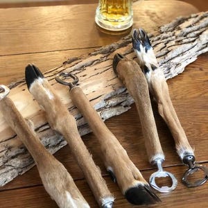 May include: A set of four bottle openers featuring handles made from preserved deer legs with dark hooves. Each handle is attached to a circular metal bottle opener mechanism. The items are arranged on a piece of rough bark.