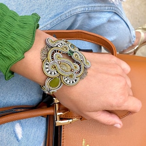 May include: A silver and gold bracelet with green and brown stones. The bracelet is made of intricate woven threads and features a unique design.