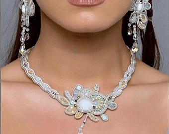 Puede incluir: Un collar de cuentas blancas y doradas con un gran colgante blanco con un diseño floral. El collar se combina con pendientes a juego.
