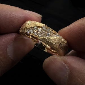 May include: A gold ring featuring a textured, rugged surface with a central channel embedded with numerous small, clear, round-cut stones. The band has a wide, irregular profile with a polished interior surface and a rough, organic exterior finish.