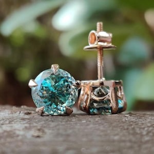 Peut inclure: Une paire de boucles d'oreilles à clous avec des pierres précieuses bleu sarcelle taille brillant rond serties dans du métal or rose. Une boucle est posée à plat pour montrer la pierre facettée, l'autre est debout, révélant la tige filetée.