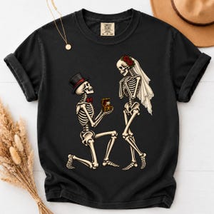 May include: A black short-sleeved t-shirt featuring a graphic of two skeletons. One skeleton wears a top hat and red bow tie while kneeling to present a ring box. The other skeleton wears a white veil with red roses. The shirt has a Comfort Colors label.