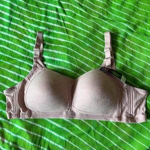May include: A light pink bra featuring molded cups with a subtle embossed floral pattern, adjustable shoulder straps, and a side panel design. A small black and white brand tag is attached to the upper edge of the right cup of the garment.