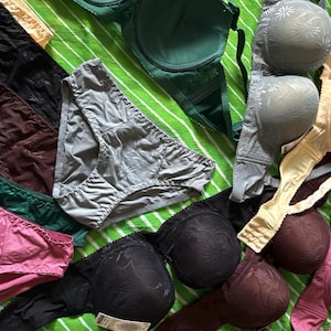 May include: A collection of assorted undergarments including several bras and panties in various colors such as black, pink, green, grey, and beige. The bras feature molded cups with a subtle floral pattern, while the panties are solid-colored.