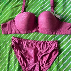 May include: A matching set consisting of a pink bra and panties. The bra features molded cups with a tonal floral pattern, adjustable shoulder straps, and a small bow at the center. The panties have a matching floral textured front panel.