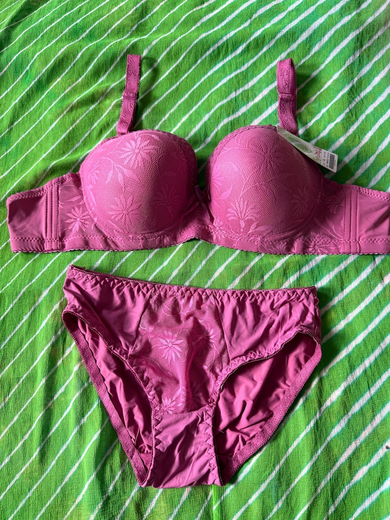 May include: A matching set consisting of a pink bra and panties. The bra features molded cups with a tonal floral pattern, adjustable shoulder straps, and a small bow at the center. The panties have a matching floral textured front panel.