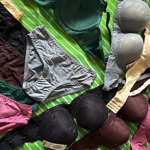 May include: A collection of assorted undergarments including several bras and panties in various colors such as black, pink, green, gray, and beige. The items feature a subtle floral pattern on the fabric, with some pieces showing adjustable straps and hook-and-eye closures.