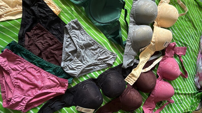 May include: A collection of assorted undergarments including several bras and panties in various colors such as black, pink, green, gray, and beige. The items feature a subtle floral pattern on the fabric, with some pieces showing adjustable straps and hook-and-eye closures.
