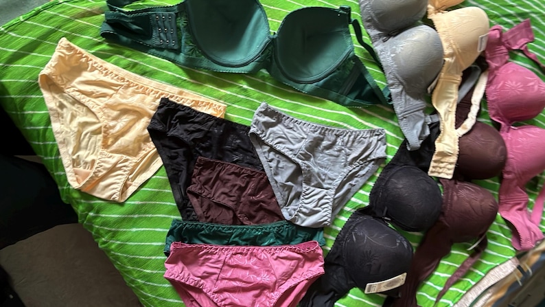 May include: A collection of assorted undergarments including several bras and panties in various colors such as green, black, gray, pink, beige, and dark brown. The items feature different textures, patterns, and styles, displayed together in a group.
