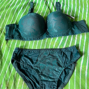 May include: A dark green lingerie set consisting of a molded cup bra and matching high-waisted briefs. Both pieces feature a tonal floral lace overlay on the front panels, adjustable shoulder straps, and a solid dark green fabric finish.