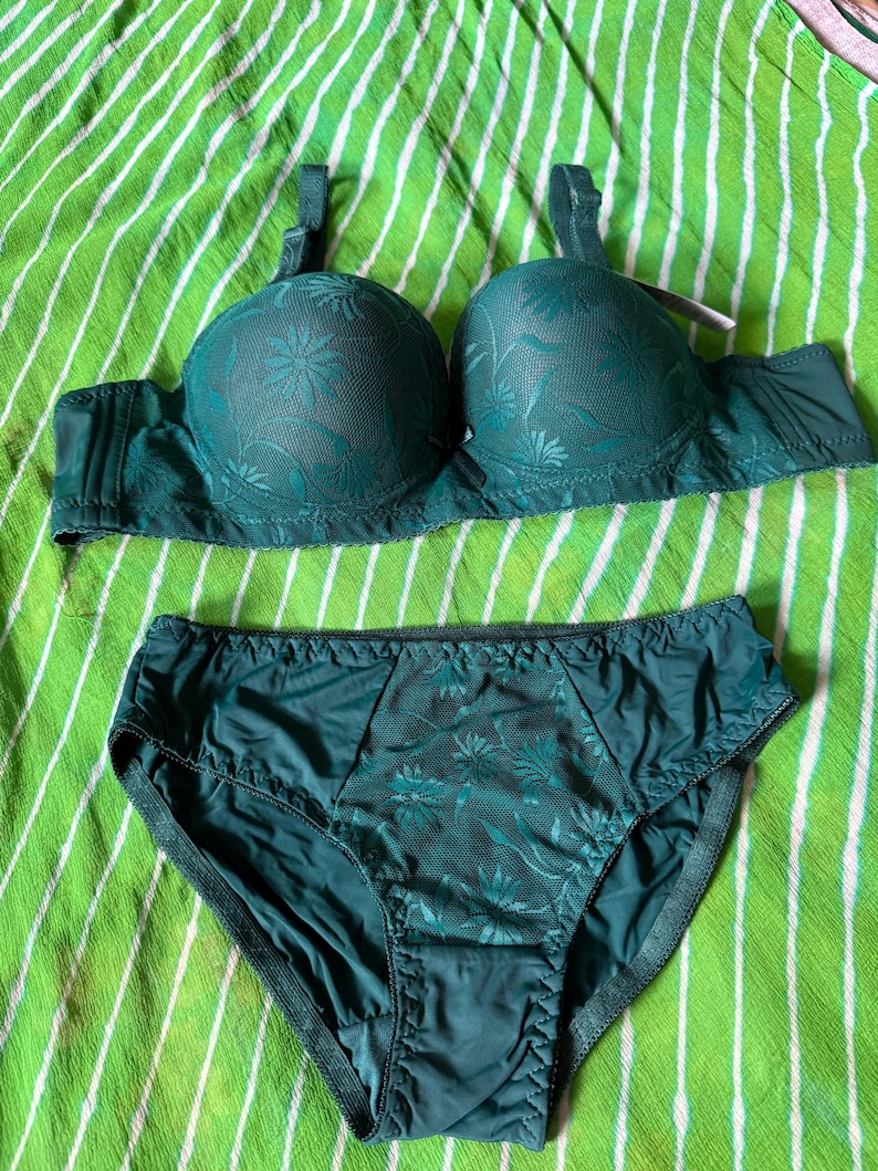 May include: A dark green lingerie set consisting of a molded cup bra and matching high-waisted briefs. Both pieces feature a tonal floral lace overlay on the front panels, adjustable shoulder straps, and a solid dark green fabric finish.