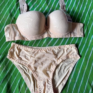May include: A beige lingerie set consisting of a molded cup bra and matching briefs. The bra features textured floral patterns on the cups and adjustable straps. The briefs have a central floral panel with smooth side sections and elastic trim.