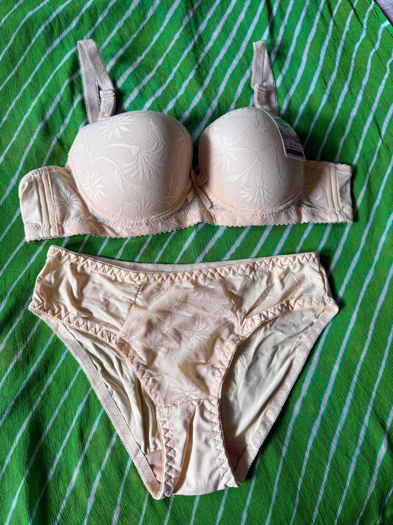 May include: A beige lingerie set consisting of a molded cup bra and matching briefs. The bra features textured floral patterns on the cups and adjustable straps. The briefs have a central floral panel with smooth side sections and elastic trim.
