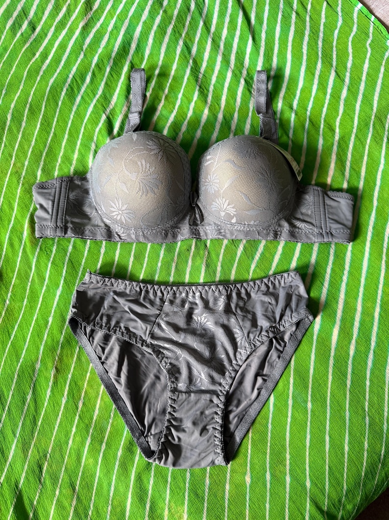 May include: A matching gray lingerie set consisting of a molded cup bra and high-cut briefs. Both pieces feature a subtle, tonal floral pattern overlaid on the fabric. The bra includes adjustable shoulder straps and a wide band, while the briefs have a smooth, solid gray back panel and a patterned front panel.
