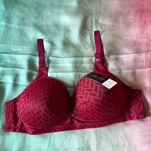 May include: A deep red bra featuring a textured geometric woven pattern across the cups and side panels. The garment includes adjustable shoulder straps and a small white tag on the right cup with the text NO. D29 SIZE 46/105 printed in black.